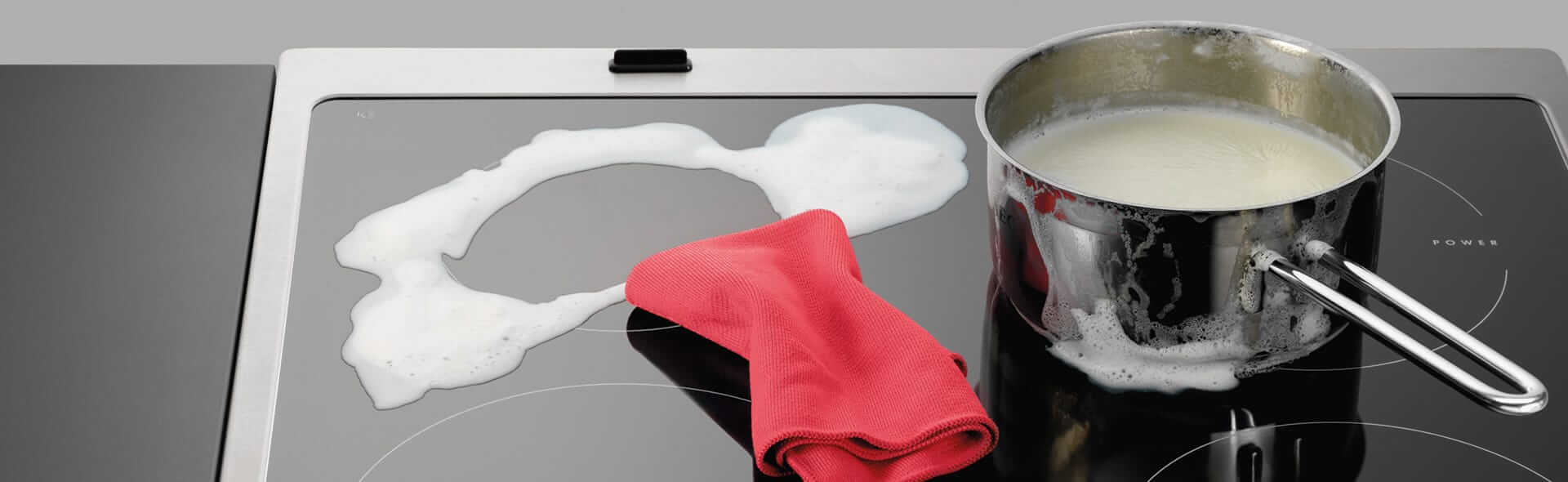 Übergekochte Milch auf Kochfeld mit Topf und rotem Tuch