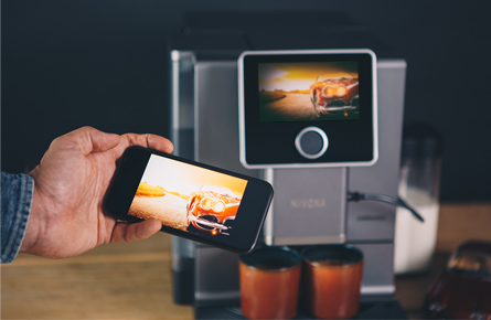 Handy zeigt gleiches Bild wie das Display der Kaffeemaschine im Hintergrund